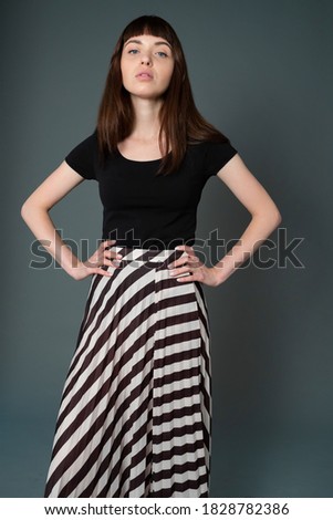 Studio portrait of a pretty brunette woman in a black t-shirt and striped skirt, standing against a plain grey background, looking at camera