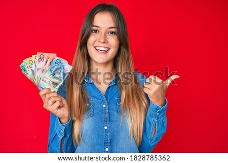 Beautiful caucasian woman holding australian dollars pointing thumb up to the side smiling happy with open mouth 