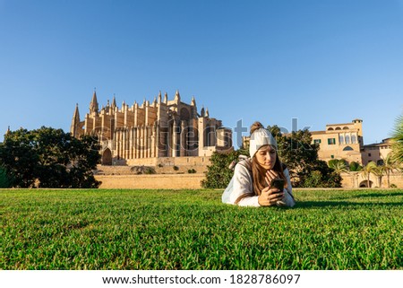 Young woman with fair skin and brown hair lying on the grass in front of the cathedral of Palma using her mobile in autumn, wears a white sweater and yarn hat with striped blue pants, clear sky