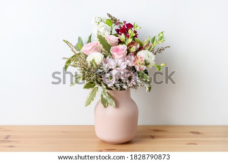 Beautiful cottage style flower arrangement, in a pink vase, on a wooden table. Flower bunch includes Roses, Snapdragons, Ranunculus, Daisy's, Chrysco, Lily of the Incas, Thryp and lush green foliage.  Royalty-Free Stock Photo #1828790873