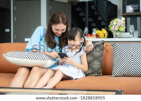 Asian cute child using a smartphone together with mother and smiling for watching video or play game while sitting on sofa in living room at home
