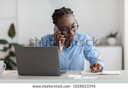 Busy Black Female Office Worker Talking On Cellphone With Client And Taking Notes, Sitting At Workplace In Modern Office, Working On Desk With Laptop, Writing Down Information To Notepad, Copy Space