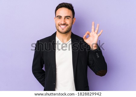 Young caucasian man isolated on purple background winks an eye and holds an okay gesture with hand.