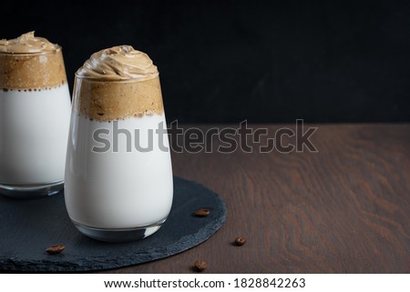 Glass of homemade dalgona drink made by whipping instant coffee powder, sugar and hot water with addition of cold milk on stony tray on dark wooden background at kitchen. Image with copy space