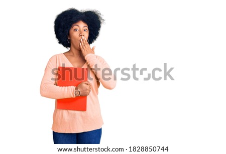Young african american girl holding book covering mouth with hand, shocked and afraid for mistake. surprised expression 