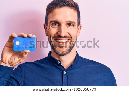 Young handsome businessman holding credit card over isolated pink background looking positive and happy standing and smiling with a confident smile showing teeth