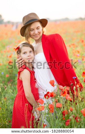 Beautiful mother and daughter in straw hats and red dresses hug each other in spring on a poppy field in the mountains