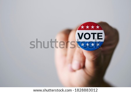 closeup of a young caucasian man with a vote badge for the United States election in his hand, on an off-white background with some blank space on the left