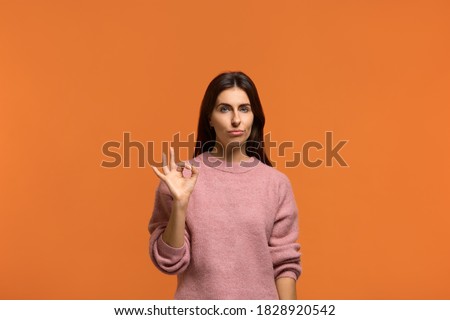 Good job but im not impressed. Not bad. Beautiful woman judging nice work show okay gesture with a neutral expression. wears in pink knitted sweater isolated on orange background