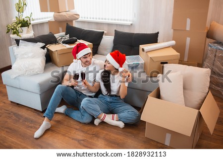 Newlywed couple celebrate Christmas or New Year in their new apartment. Young happy man and woman drinking wine, celebrating moving to new home and sitting among boxes.