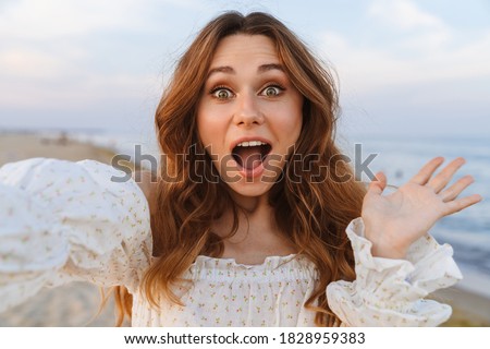 Young caucasian excited brunette woman in dress wondering at camera and taking selfie on beach