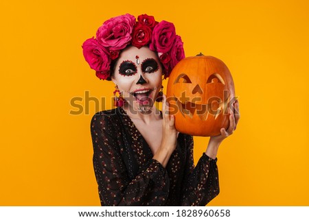 Excited caucasian girl in halloween makeup posing with pumpkin isolated over yellow background