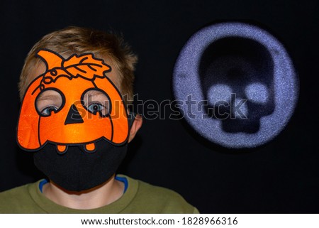 Front view close up Halloween decorations in new reality of COVID-19 pandemic. boy in orange pumpkin and medical mask.Moon reflector with human skull silhouette on dark background