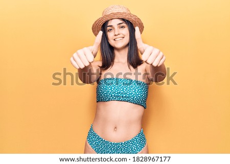 Young beautiful girl wearing bikini and summer hat approving doing positive gesture with hand, thumbs up smiling and happy for success. winner gesture. 