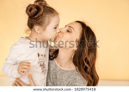 Close-up of a little girl in a white sweater with a beautiful hairstyle who is kissed by her mother, in the background is a yellow monochrome background. Part and love concept. Down Syndrome.