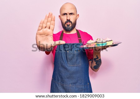 Young handsome man wearing apron holding cupcake with open hand doing stop sign with serious and confident expression, defense gesture 