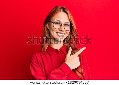 Young beautiful redhead woman wearing casual clothes and glasses over red background cheerful with a smile of face pointing with hand and finger up to the side with happy and natural expression