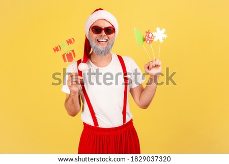 Happy positive gray bearded man in santa claus costume holding paper cards on sticks, celebrating christmas and congratulating with new year. Indoor studio shot isolated on yellow background