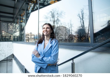 Cheerful woman wearing blue formal suit standing outside office windows on background looking up confident and smiling hopeful, arms crossed in confidence for the future