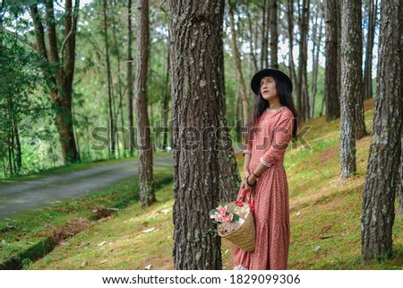 A beautiful woman standing beside a pine tree with a basket of white flowers in hand in the park.