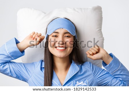 Close-up of happy and pleased smiling asian girl in blue pajamas and sleeping mask lying in bed on pillow, stretching satisfied and grinning after having good night sleep, waking up energized