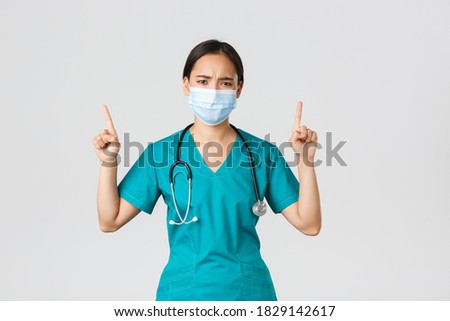 Covid-19, coronavirus disease, healthcare workers concept. Gloomy disappointed asian nurse, physician in medical mask and scrubs, pointing fingers up and frowning perplexed, look frustrated