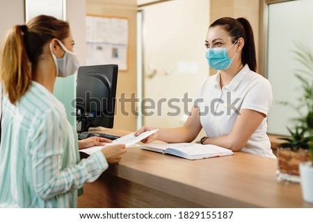 Female receptionist taking application form from her customer at health spa check in counter. They are wearing protective face masks due to COVID-19 pandemic.  Royalty-Free Stock Photo #1829155187