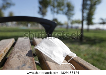 Facial mask is left on the chair or bench in park It is on wood It becomes risky to transmit or spread the virus Weather is warm in summer or spring and sunny Green grass and trees are in garden