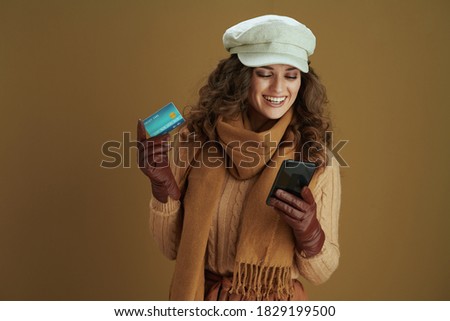 Hello october. smiling trendy housewife in sweater with smartphone, leather gloves and credit card buying on internet isolated on bronze background.