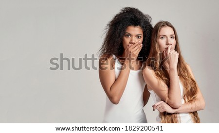 Portrait of two young diverse women in white shirts looking shocked, pensive at camera, covering mouth with hand while posing together isolated over grey background. Diversity, friendship concept