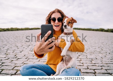 A beautiful woman sitting in a park with her cute little dog. She is taking a self-portrait photo with her dog with the phone. Technology lifestyle with pets