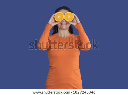 Brunette woman with oranges on her face, wearing orange t-shirt isolated on a pantene blue background