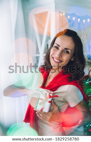 Portrait of smiling beautiful young woman posing indoors with a gift box in hands. Christmas tree on the background. Vertical shot.