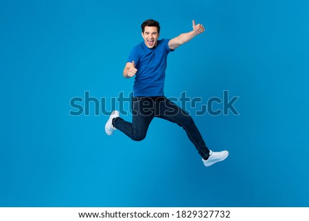 Smiling handsome American man joyfully jumping and doing double thumbs up gesture isolated on blue studio background