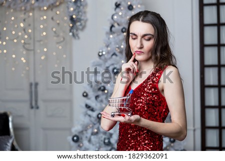 A young woman in a red dress thought about shopping for gifts, holding in her hand a small shopping cart against the background of the New Year tree.
