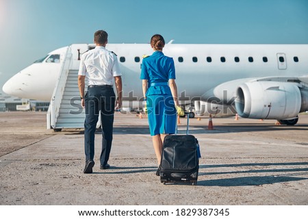 Man and woman in aviation uniform are walking to plane stairway with luggage before departure Royalty-Free Stock Photo #1829387345