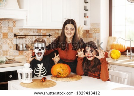 Happy kids celebrate halloween in kitchen at home in costumes and make-up with pumpkin lantern Jack looking at camera and smiling
