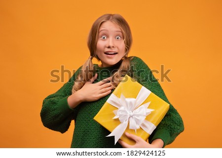 Image of charming blonde girl 12-14 years old in warm green sweater smiling and holding present box with white bow. Studio shot, yellow background. New Year Women's Day Birthday Holiday concept