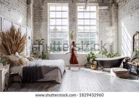 Silhouette of young woman walking at home with interior design in bohemian style against loft brick wall with large windows. Dreamy female standing between cozy bed in bedroom and bathroom bath