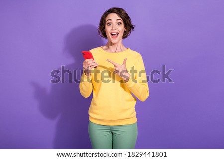 Photo of crazy girl point index finger smartphone scream wear yellow sweater green pants isolated on purple color background