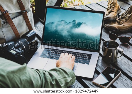 Traveler or travel photographer working on laptop computer outdoor among his equipment in the background (backpack, camera, smartphone, boots). Mountain landscape photo on the screen. Royalty-Free Stock Photo #1829472449