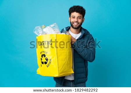 Young Moroccan man holding a bag full of plastic bottles to recycle over isolated background pointing to the side to present a product