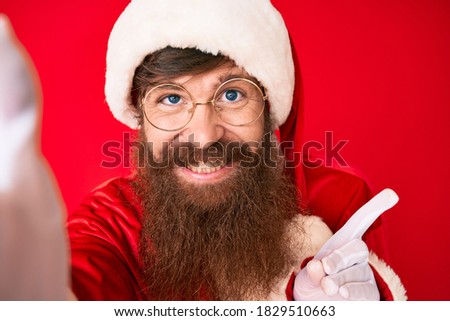 Handsome young red head man with long beard wearing santa claus costume taking a selfie smiling happy pointing with hand and finger to the side 