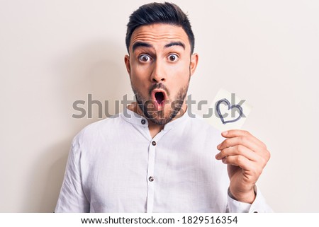 Young handsome romantic man with beard holding reminder with heart shape symbol scared and amazed with open mouth for surprise, disbelief face