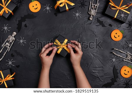 Female hands decorate a Halloween gift surrounded by boxes and decorations on a black background. Festive set with funny pumpkins, bats, skeletons and spiders.