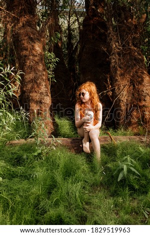 A red-haired girl in a bathing suit sits thoughtfully among the greenery of the forest.