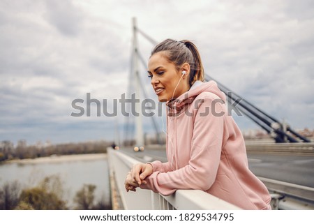Beautiful young sportswoman leaning on the bridge railing, resting, listening music and looking at river. Urban life concept.