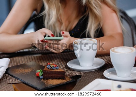 Young blond woman takes pictures of food on a smartphone at a table in a cafe. Cups with coffee and plates with cake. Selective focus.