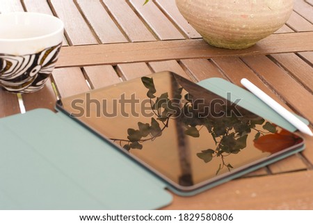 A tablet with a blue cover  and a cup of coffee or tea on a brown wooden table in a backyard. clay pot with a green plant in it. Green trees and lawn on the background.