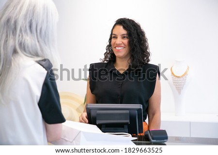 Happy positive shop cashier talking to customer and laughing at checkout. Medium shot. Shopping concept